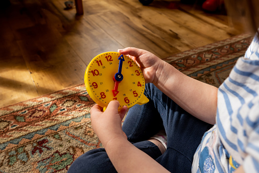 A young girl sitting cross legged on a rug covered wooden floor. The girl is practicing telling the time using a yellow plastic toy clock face, with blue and red hands which can be moved into different positions, with 12 and 24 hour numbers marked on the circular teaching toy.