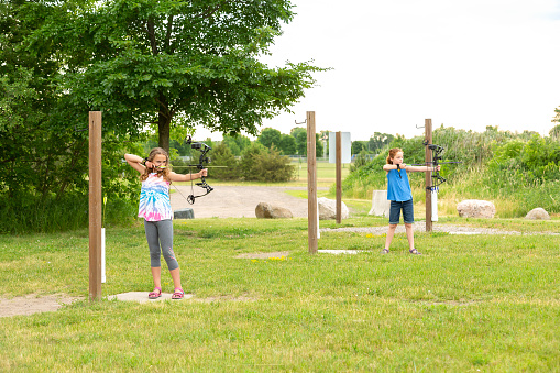 Two young girls (sisters) practicing archery at an outdoor range on a summer day.