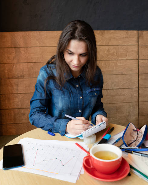 Girl in a blue denim shirt sitting in a cafe makes notes in a notepad with a pen. A smartphone, sheets with various charts, glasses in a case, a red mug with green tea, felt-tip pens are on the table una chica con una camisa de mezclilla azul sentada en un café hace notas en un bloc de notas con un bolígrafo. un teléfono inteligente, sábanas con varios gráficos, gafas en una caja, una taza roja con té verde, rotuladores están en la mesa