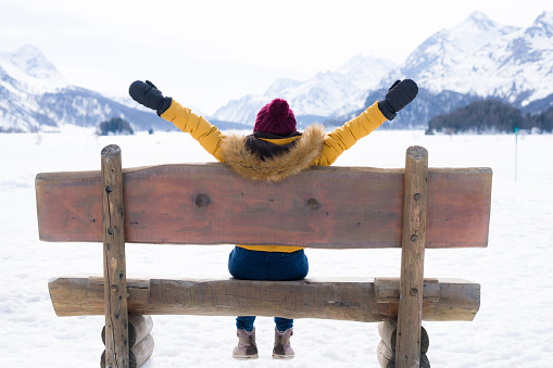 amazing Swiss Alps viewpoint from wooden bench - happy woman sitting on bench looking snow mountain landscape in front of frozen lake in Switzerland on Winter