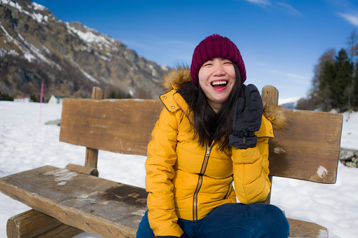 young happy and beautiful Asian Chinese woman in yellow jacket and winter hat smiling cheerful on bench over frozen lake covered in snow in Swiss Alps mountains