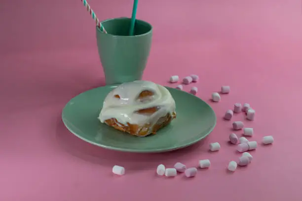 a delicious cinnamon-sugar spiral bun with cream on a green plate with a mug of the same color on a pink background with sugar clouds