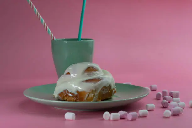 a delicious cinnamon-sugar spiral bun with cream on a green plate with a mug of the same color on a pink background with sugar clouds