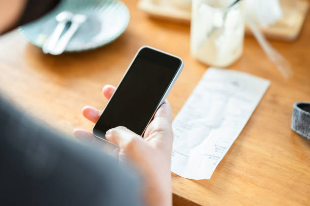 Use Mobile Phone with Food Bill on the Table Closeup woman hand use the smart phone after meal in the coffee shop, with bill of food and drink on the table, scan or online payment, bill calculating concept receipt stock pictures, royalty-free photos & images
