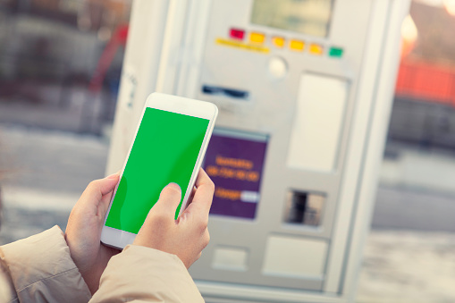 Closeup of a person using a mobile phone with green screen next to a parking ticket machine at a parking lot.