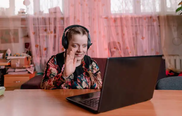Photo of a disabled girl with slanted eyes sits at a table with headphones on and communicates while looking at a laptop. training of people with disabilities