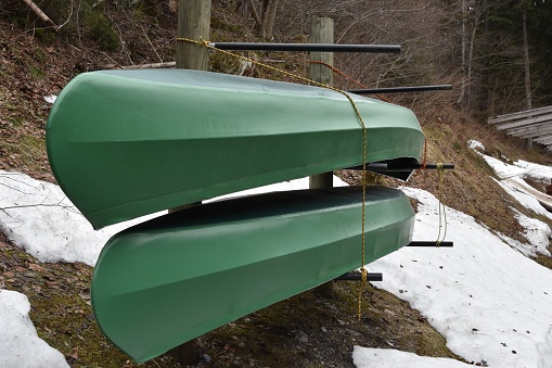 Two green kayaks  stored in racks upside down in side view. They wait for start of water sport season. There are patches of snow around them.