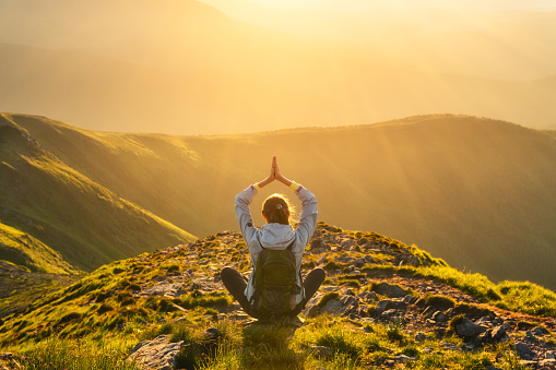 Young woman with backpack sitting on the mountain peak and beautiful mountains in fog at sunset in summer. Landscape with sporty girl, green forest, hills, sky, sunbeams. Travel and tourism. Yoga