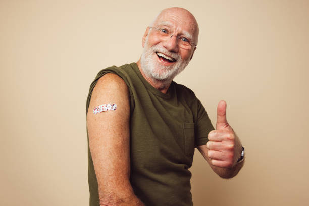 I got the immunity vaccine Portrait of a senior male smiling and showing thumbs up after getting a vaccine. Mature man with white beard sitting against brown background feeling positive after receiving immunity vaccination. flu vaccine photos stock pictures, royalty-free photos & images