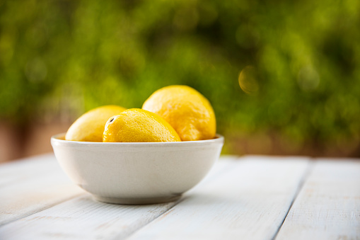 This is a close up photograph with a group of fresh lemons in a bowl on a white picnic bench with a tree in the background out of focus