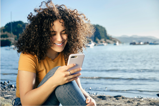 Attractive curly hair happy woman sitting on the lakeshore using a smart phone to read or write a message. Vacation concept and technology
