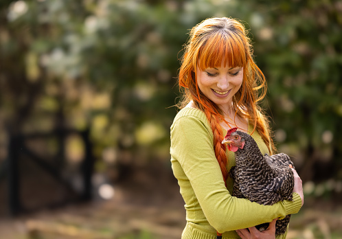 Young 30 year old woman with red/orange hair, alone on a farm, holding a pet chicken