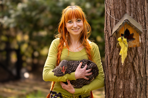 Young 30 year old woman with red/orange hair, alone on a farm, holding a pet chicken