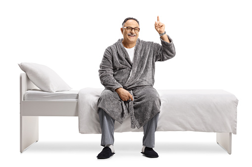 Smiling mature man in pajamas and a robe sitting on a bed and pointing up isolated on white background