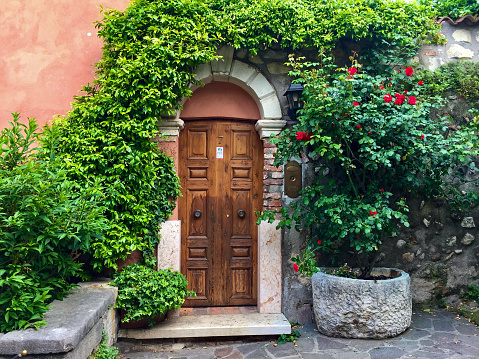 Fragment of facade with wooden door, pink wall decorated with ivy. Red rose in stone wase. Old house stone wall, cobblestone. Traditional Italian house