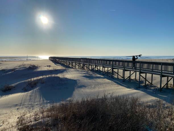 Surfer walking down a boardwalk at Assateaque Island in Maryland, USA Surfer walking down a boardwalk that connects to the beach at Assateaque Island in Maryland, USA. The morning sun is bright as it rises behind him casting him in silhouette. eastern shore sand sand dune beach stock pictures, royalty-free photos & images