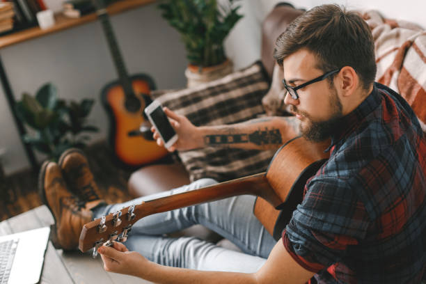 istock Young guitarist hipster at home with guitar tuning using app 1312386268