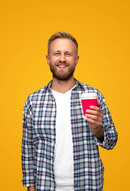 Bearded guy with drink to go Positive bearded man in checkered shirt smiling for camera and carrying takeaway cup of hot beverage in morning against yellow background single cup stock pictures, royalty-free photos & images