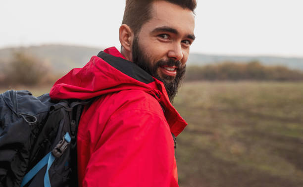 Side view of positive adult bearded male hiker in red outerwear and with backpack looking at camera during travel through autumn nature