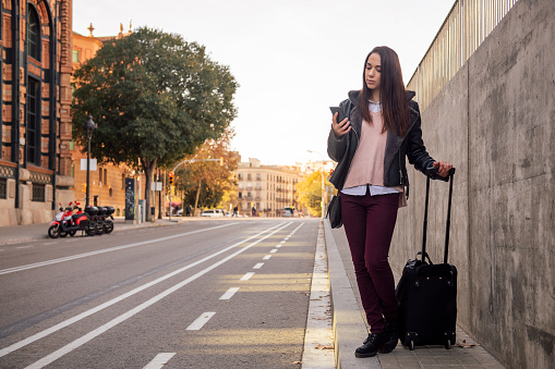 young woman with suitcase waiting for a taxi on the street while consulting her phone, concept of travel and technology, copyspace for text