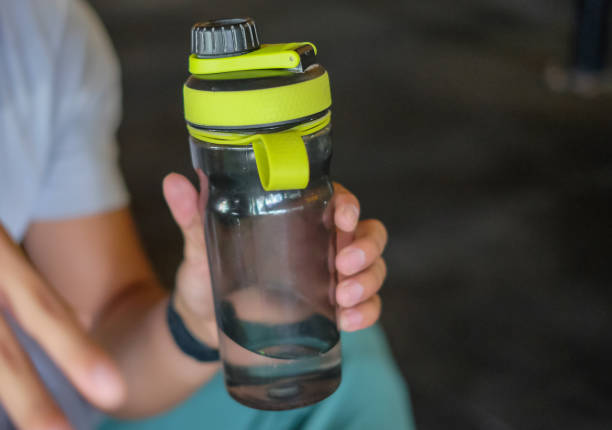 Close-up shot of an unrecognizable male hands holding a reusable water bottle plastic. He's sitting on the ground after finishing his exercise in a gym.