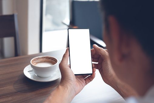 Mockup image of man hand holding and watching blank screen mobile smart phone, reading news via application with cup of coffee on wooden table in loft coffee shop, over shoulder view