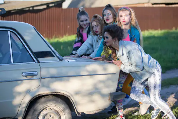 Photo of Young cheerful girls are pushing an old car. Women in the style of the 90s.