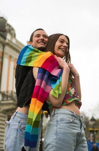 A young lesbian couple embraces their love. They are hugging on the street covered together with a rainbow colors scarf.