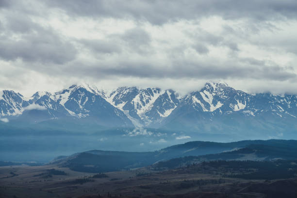 istock Scenic mountain landscape with great snowy mountain range among low clouds and green forest in valley at early morning. Atmospheric alpine scenery with blue white high mountain ridge under cloudy sky. 1312414805