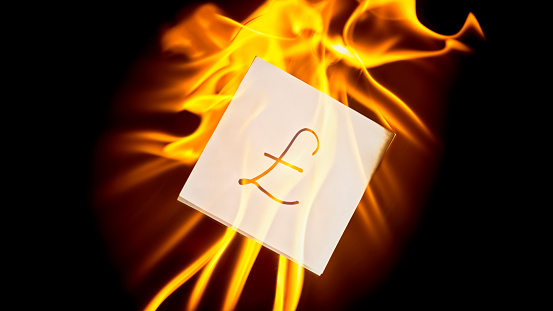 Close-up of white piece of paper with pound Sterling sign burning in flames against black background.