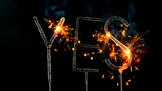 Close-up of sparklers shaped to present the word 'YES' burning and emitting sparks against black background.
