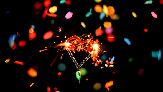 Close-up of heart shaped sparkler emitting sparks while confetti falling against black background.