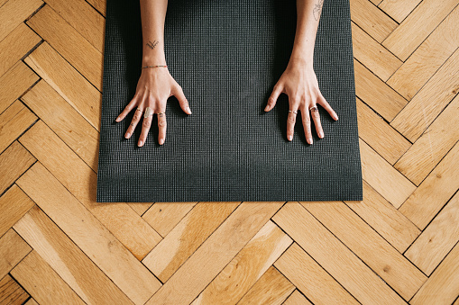 A pair of hands with rings is resting on a dark yoga mat. Wooden parquet harringbone pattern is in contrast with the mat. A no-face horizontal photo with copy space.