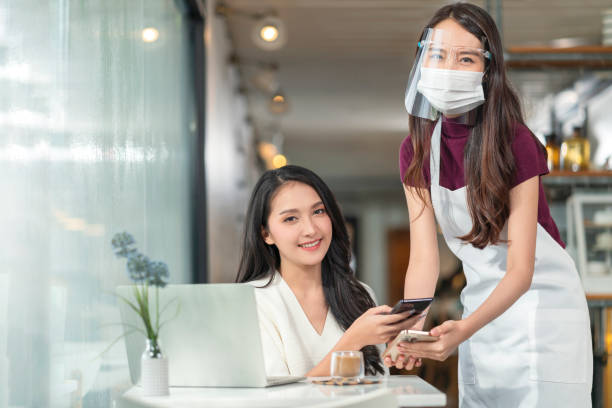 istock Contactless payment with mobile in restaurant. Waitress standing, wearing protective face mask and apron with asian smiling customer with her smartphone on hand about to pay by qr code payment system 1312439860
