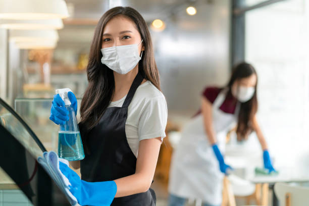 atraente positivo jovem asiático pessoal em avental uniforme pulverizando detergente ou álcool no armário de produto de balcão limpando-o com pano macio ao desinfetar café na manhã segura e limpa - cleaning services - fotografias e filmes do acervo