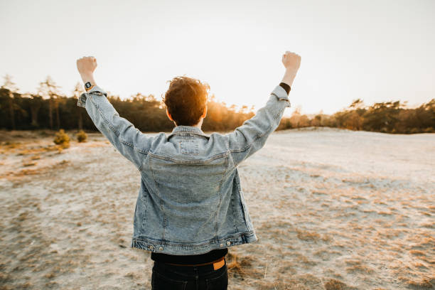 person with raised fists from behind person with raised fists from behind short brown hair wearing jeans jacket at sunset in frint of a sandy area denim jacket stock pictures, royalty-free photos & images