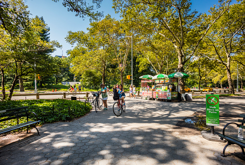New York City, United States - August 24, 2017: In Central Park, kiosk of a hot dog and ice cream sidewalk vendor, with people walking and women with bicycle.