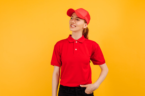 Smiling young woman courier wearing red uniform posing on yellow studio background, delivery concept
