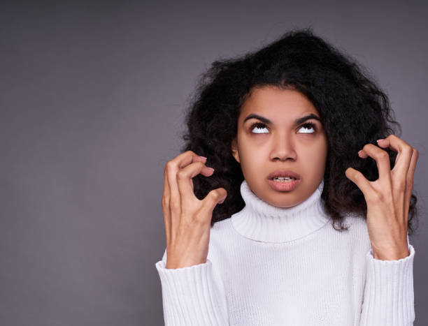 Angry, aggressive dark-skinned young girl on a gray background. Aggressive young dark-skinned girl, in a white sweater, with magnificent curly hair, shows a grimace of disgust, hatred, raised her hands to her face and looks up. anger stock pictures, royalty-free photos & images