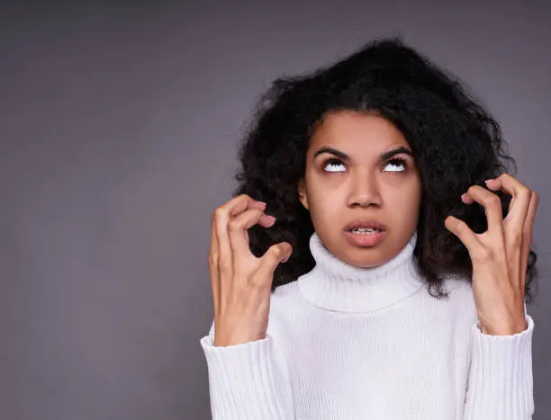 Aggressive young dark-skinned girl, in a white sweater, with magnificent curly hair, shows a grimace of disgust, hatred, raised her hands to her face and looks up.