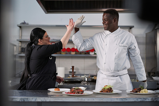 Cheerful chef assistance, serving dish, while her chef Jamaican ethnicity observing and helping her