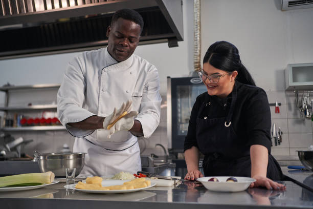 Excited female chef assistant learning new kneading technique from the professional Professional male chef, Jamaican ethnicity, kneading and preparing the dough in the modern commercial restaurant kitchen while his female assistant observing him cooking show stock pictures, royalty-free photos & images