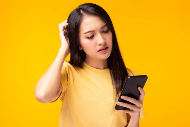 hermosa mujer joven se confunden y frustran de las compras de aplicaciones en línea chica sostener el teléfono móvil, mirar el teléfono celular con la cara de expresión ella se decepciona de teléfono roto fondo amarillo - people worried confusion women fotografías e imágenes de stock