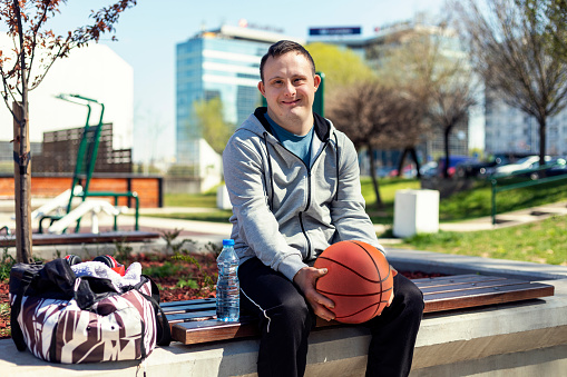 Photo of happy young man with down syndrome on basketball court in public park. He sitting on the bench with basketball ball in his hands.