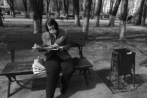 Moscow region, Russia - April 14, 2021: Senior woman is reading newspapers on park bench