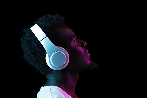 Close-up young African-American man in headphones isolated on dark background in multicolored neon light. Concept of human emotion, facial expression, youth culture. Copy space for ad. Side view
