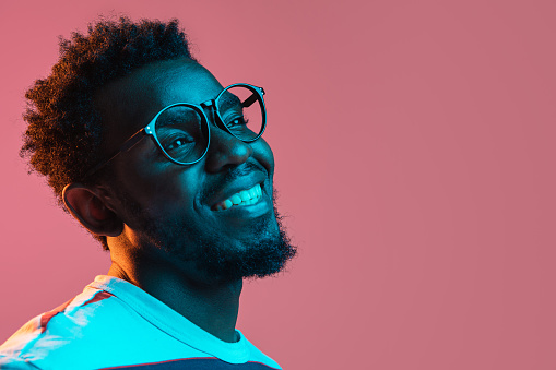 Laughing. Close-up young African-American man in glasses isolated on pink background in multicolored neon light. Concept of human emotion, facial expression, youth culture. Copy space for ad