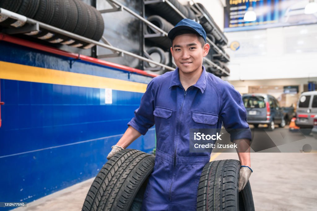 Young auto mechanic at work Car mechanic holding car tires in the auto repair shop. Young male apprentice at work, smiling at camera. Auto Mechanic Stock Photo