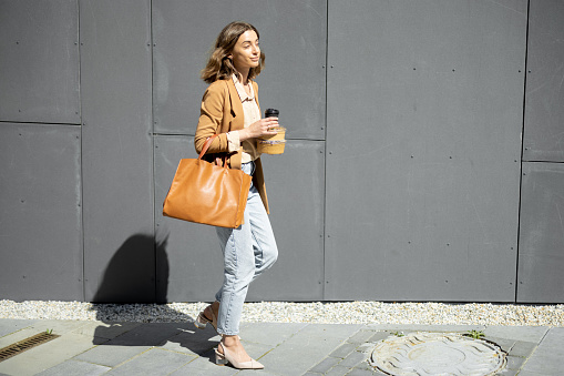 Pretty woman walking near wall of the office building with takeaway food and coffee. Healthy meal for workers. Lifestyle casual clothing.