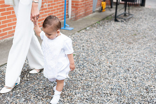 Small baby from 1 to 2 years old walks holding the hand of his mother wearing his christening gown heading to church